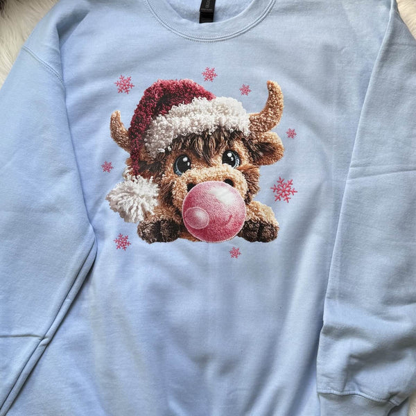 Light blue sweatshirt with a cartoon cow wearing a Santa hat and blowing a pink bubble, surrounded by snowflakes.