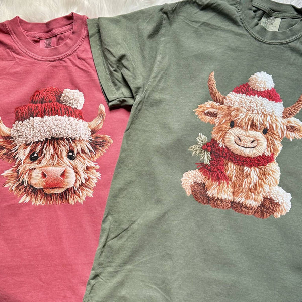 Two shirts, one red and one green, featuring cartoon Highland cows with festive santa hats and scarves.