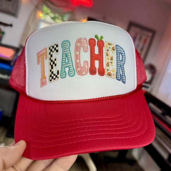 Red and white cap with 'TEACHER' text in colorful letters held in a hand.