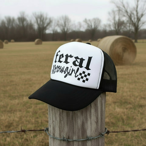 Black and white trucker hat with “Feral Cowgirl” text and checker accents on a white foam front panel.