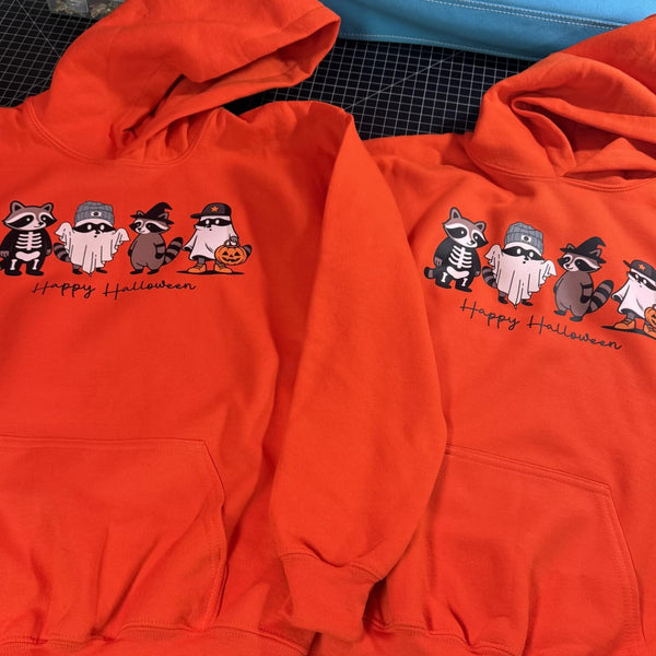 Two orange hoodies with Halloween-themed raccoon designs on a checkered surface.