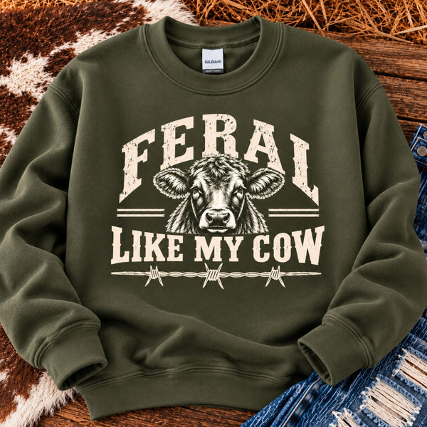 Military green Gildan Feral Like My Cow sweatshirt with vintage distressed cattle graphic, displayed flat with blue jeans and cowhide on a rustic wooden background.