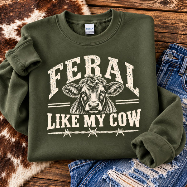 Feral Like My Cow military green crewneck sweatshirt with antique white cow graphic and barbed wire design, folded beside distressed jeans on rustic wood.
