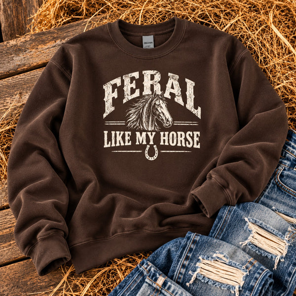 Feral Like My Horse Western graphic sweatshirt in chocolate brown with distressed horse illustration and horseshoe detail, displayed beside ripped blue jeans.