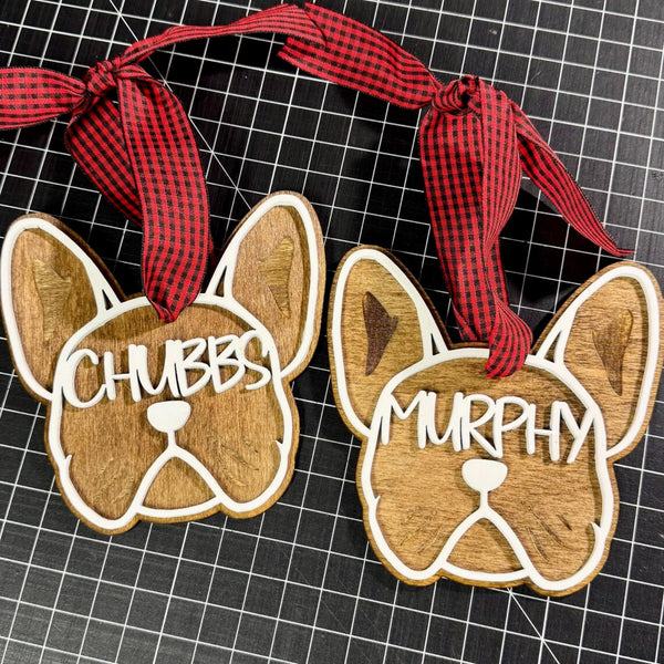 Two personalized wooden French Bulldog ornaments with red and black gingham ribbon hangers, displayed on a black grid cutting mat. One ornament is engraved with 'CHUBBS' and the other with 'MURPHY'.