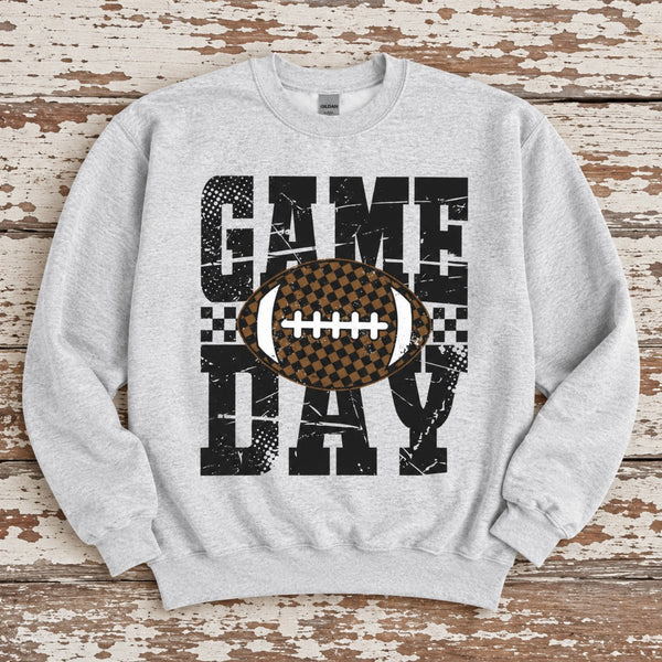 A light heather gray crewneck sweatshirt laid flat on a rustic, distressed wooden background. The sweatshirt features a large graphic print reading 'GAME DAY' in distressed black block letters, with a brown and black checkered football graphic in the center.
