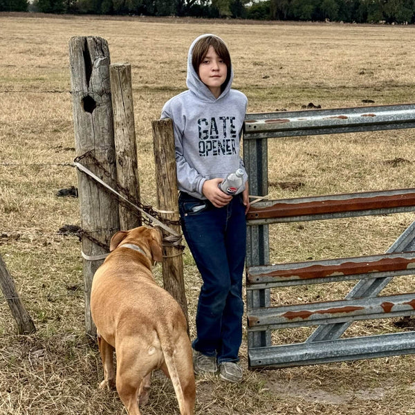 Boy in a field with a dog standing next to a gate wearing a gray Gate Opener hoodie with cattle branding.