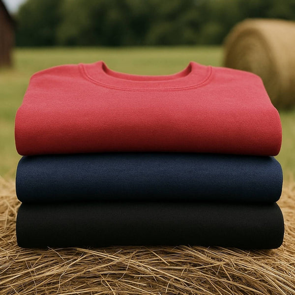 Red, navy, and black fleece-lined pullover sweatshirts folded on a hay bale in a rustic country setting.