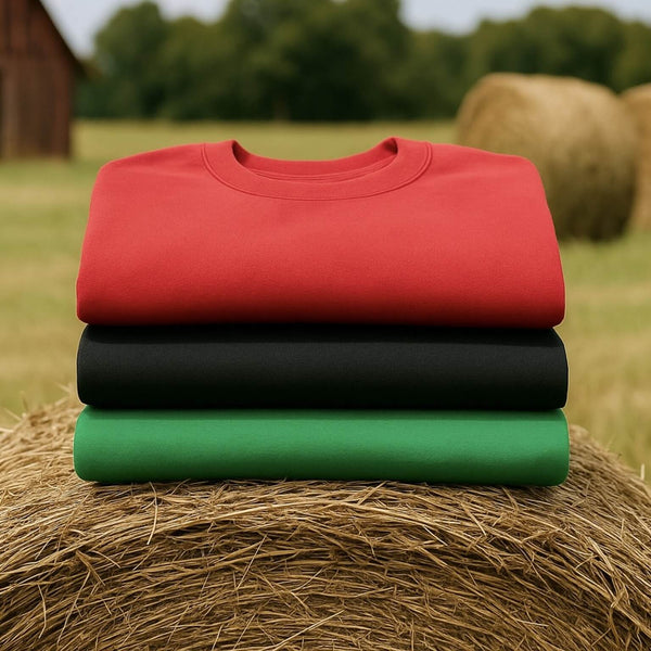 Red, black, and green fleece pullover sweatshirts folded on a hay bale with barn and hay rolls in the background.