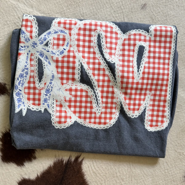 USA graphic tee featuring red gingham patchwork lettering with lace trim and blue floral bow on Comfort Colors Denim Blue shirt