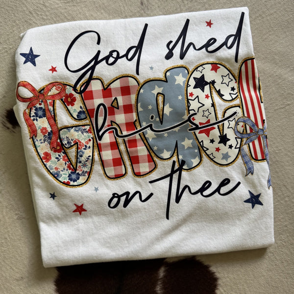 God Shed His Grace on Thee patriotic graphic tee featuring red white and blue patchwork lettering and bow accents on white shirt