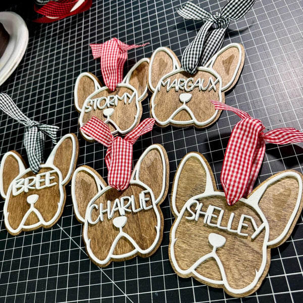 Five personalized wooden French Bulldog ornaments with white painted outlines, red and black gingham ribbon hangers, and custom names (Bree, Stormy, Margaux, Charlie, Shelley), displayed on a black gridded cutting mat.