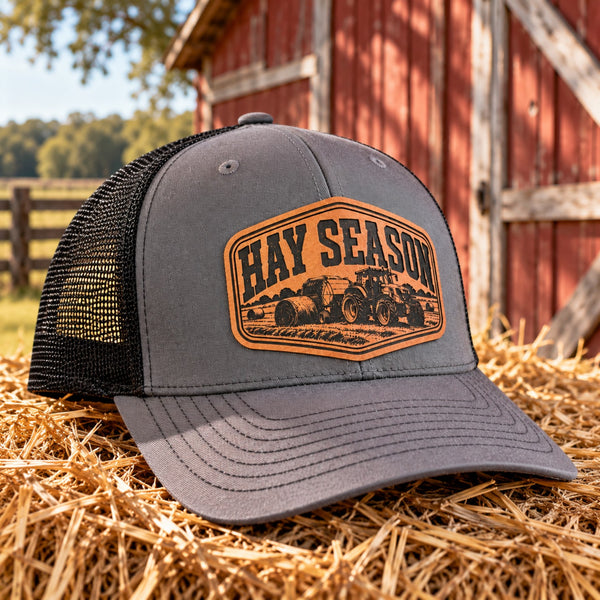 Gray and black Hay Season farm trucker hat with tractor leather patch beside a red barn