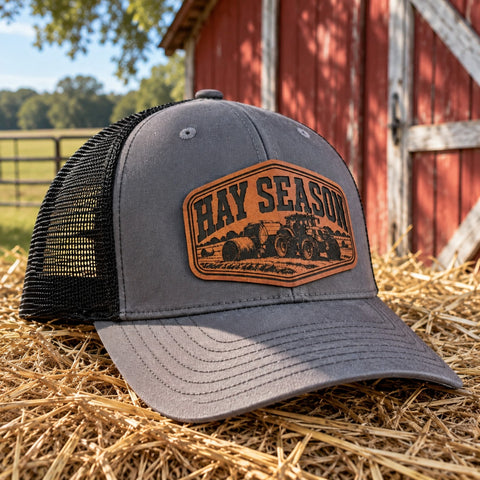 Gray and black Hay Season farm trucker hat with tractor leather patch beside a red barn