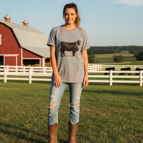 Young woman wearing a gray t-shirt with a black cow graphic and turquoise necklaces standing outside by a barn.