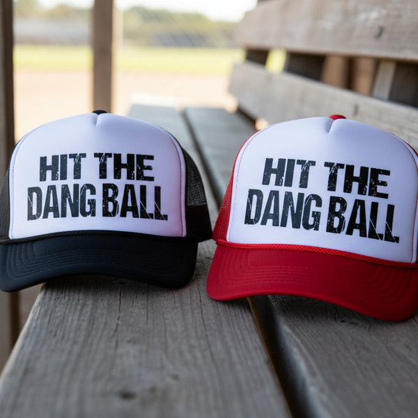 Black and red “Hit The Dang Ball” foam trucker hats displayed on wooden baseball bleachers with distressed sporty lettering for baseball and softball game day outfits
