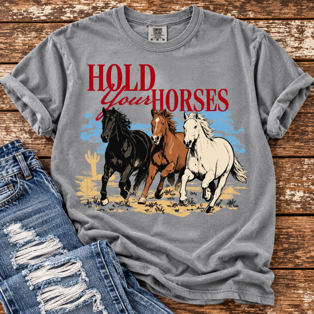 Gray Comfort Colors Hold Your Horses graphic tee with three galloping horses and red Western lettering, styled with distressed blue jeans on rustic wood.