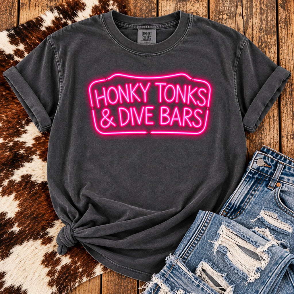 Honky Tonks & Dive Bars graphic tee with hot pink neon lettering on Pepper Comfort Colors shirt styled with distressed jeans and cowhide.
