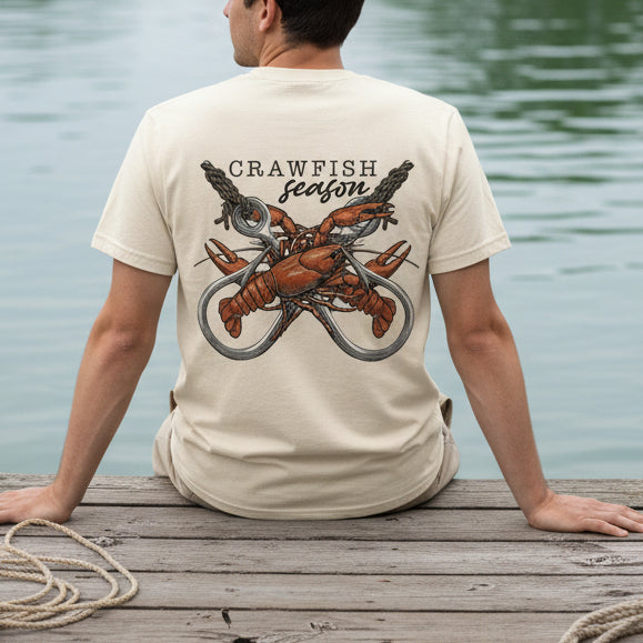 Man wearing ivory comfort colors t-shirt with crawfish graphic and 'Crawfish Season' text sitting on a fishing dock