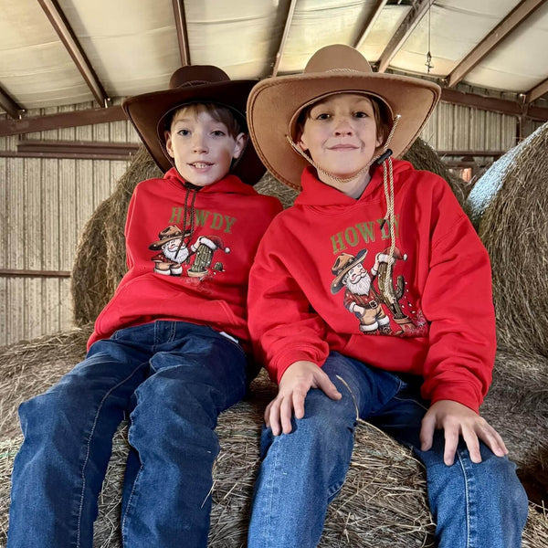 Two children wearing red hoodies with cowboy santa graphics sitting on hay bales.