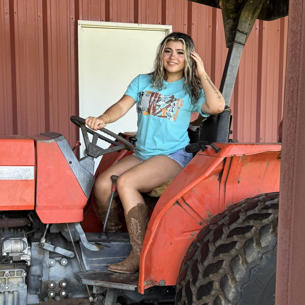 Woman sitting on a red tractor wearing a blue cowgirl t-shirt with a howdy graphic design.