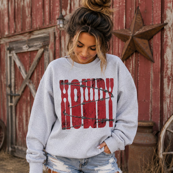 Young woman looking down while wearing a Howdy barbed wire graphic sweatshirt in Ash Gray, standing in front of a rustic red barn with distressed denim shorts and a Western country backdrop.