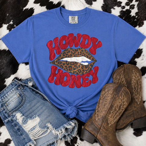 A flo blue Comfort Colors graphic T-shirt laid flat on a black-and-white cowhide background, styled with distressed denim shorts and brown cowboy boots. The shirt has a large red 'HOWDY HONEY' text graphic over a lip illustration with leopard print and white teeth.