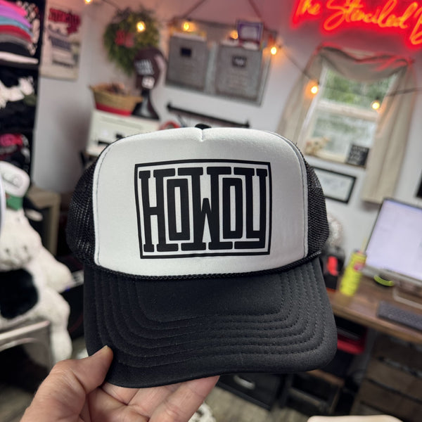 Black and white cap with 'HOWDY' logo held in front of a casual indoor setting.