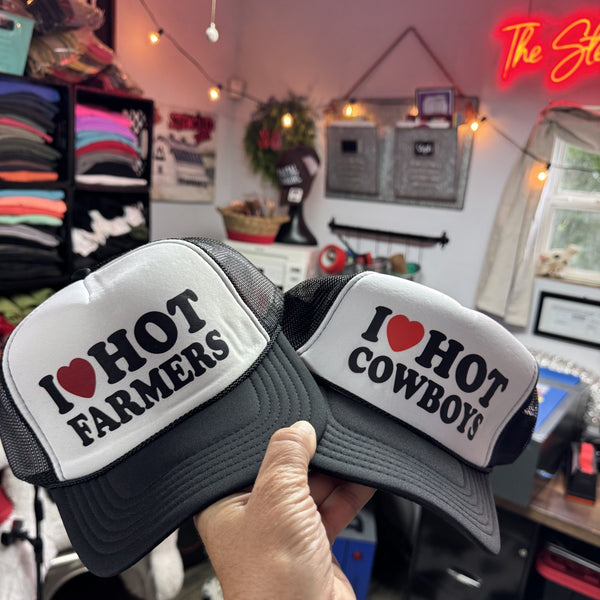 Two hats with 'I ❤️ HOT FARMERS' and 'I ❤️ HOT COWBOYS' text held in a store setting black and white foam trucker hats ready to ship from The Stenciled Barn Barn Sale.