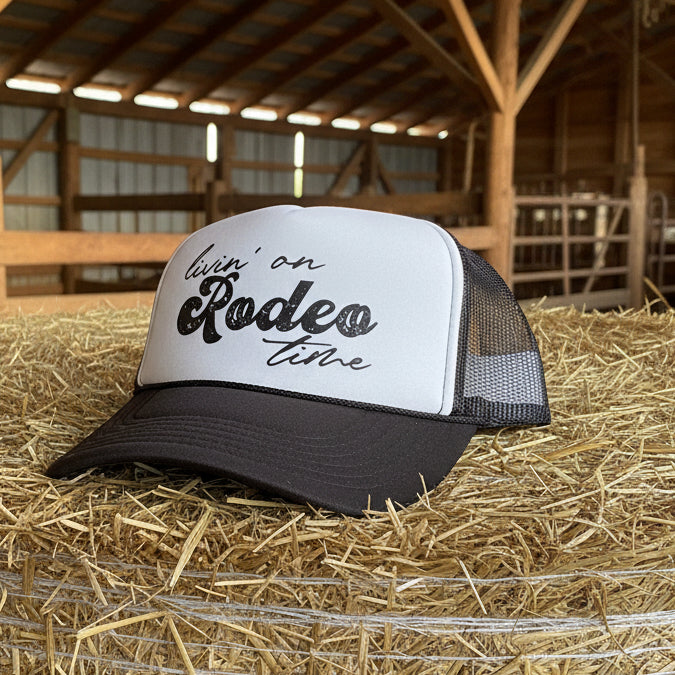 Trucker hat with 'livin' on Rodeo time' text sitting on a haystack in a barn setting.