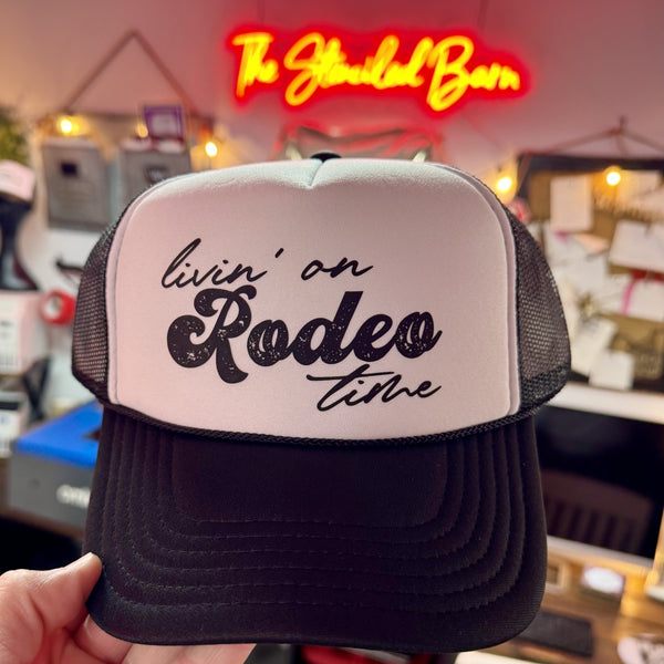 Trucker hat with 'livin' on Rodeo time' text held in front of a neon sign.