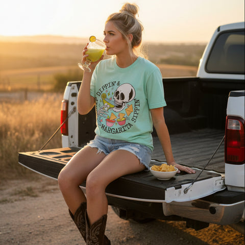 Woman wearing a light green cinco de mayo graphic tee drinking a lime margarita and eating chips, sitting on a white truck tailgate 