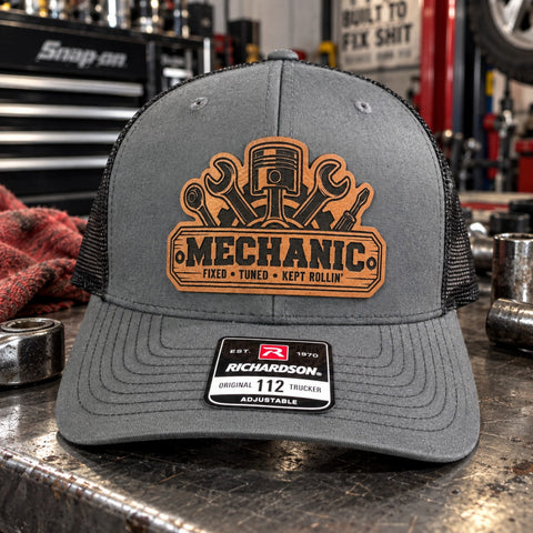 Gray Mechanic leather patch trucker hat displayed on a rustic mechanic toolbox