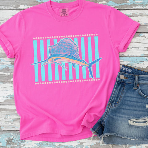 Neon pink Comfort Colors graphic tee featuring a pastel sailfish design with turquoise striped background on a white wooden backdrop