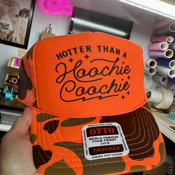 Woman holding neon orange camo trucker hat with Hotter Than A Hoochie Coochie graphic design in rustic country workshop setting