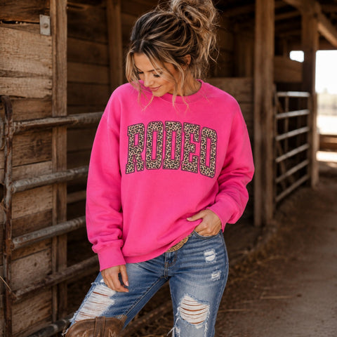 Woman wearing hot pink leopard Rodeo sweatshirt with ripped jeans and cowboy boots in a rustic barn