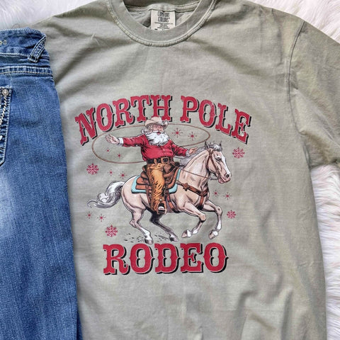 Green t-shirt with Santa riding a horse and 'North Pole Rodeo' text, next to blue jeans.
