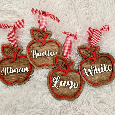 Four personalized wooden apple ornaments with red borders, red-and-white gingham ribbon hangers, and white script names, arranged on a white faux-fur surface.