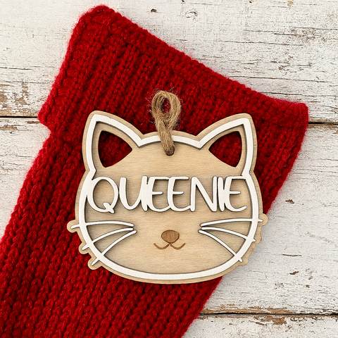 Wooden ornament shaped like a cat's face with 'QUEENIE' on it, placed on a red knitted fabric.