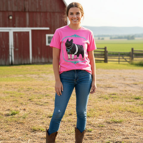 Woman wearing neon pink comfort colors brand t-shirt with a graphic of a pig wearing a turquoise necklace standing by a red barn.
