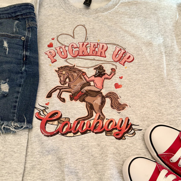 Gray t-shirt with 'Pucker Up Cowboy' graphic design, jeans, and red sneakers on a light background