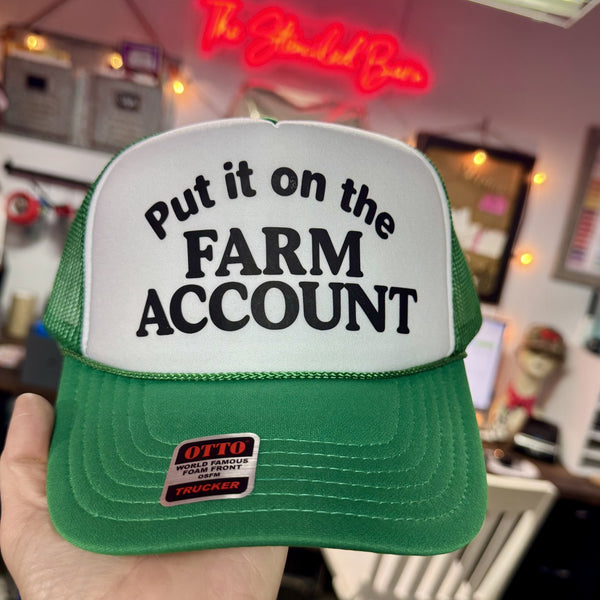 Green and white cap with 'Put it on the Farm Account' text held in front of a neon sign.
