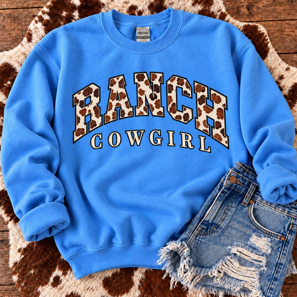 Ranch Cowgirl blue sweatshirt with brown cow print lettering styled on cowhide rug with distressed denim shorts