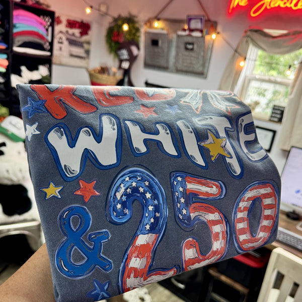 Rustic patriotic “Red White & 250” graphic tee displayed on denim blue Comfort Colors brand t-shirt with printed Americana lettering, red white and blue stars, and vintage USA-inspired design details in a cozy Southern boutique setting.