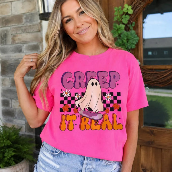 A woman wearing a bright pink graphic t-shirt with a 'CREEP IT REAL' Halloween ghost design, paired with light-wash denim jeans, standing in front of a rustic stone and wood door background.