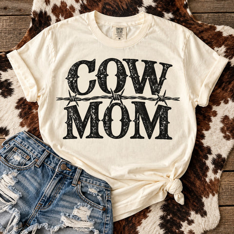 Ivory Comfort Colors Cow Mom graphic tee with distressed black western lettering and barbed wire design, styled with ripped denim shorts on rustic cowhide.