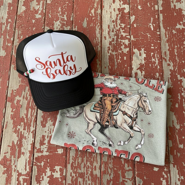 Santa Baby trucker hat and cowboy Santa Christmas graphic tee on white faux fur background – rustic western holiday apparel set from The Stenciled Barn.