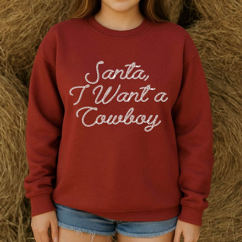 Woman wearing a rustic red Christmas pullover that says “Santa, I Want a Cowboy” in white rope-style lettering, western holiday apparel.