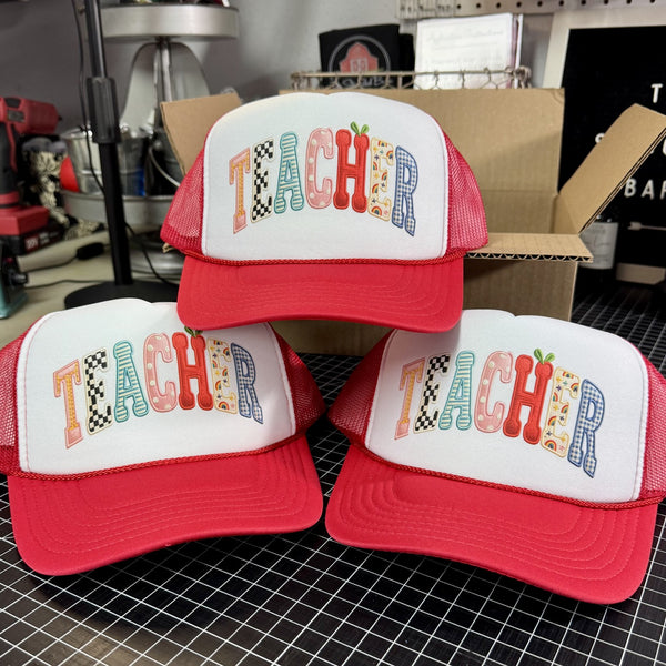 Three 'TEACHER' trucker hats with colorful text on a work table surface.