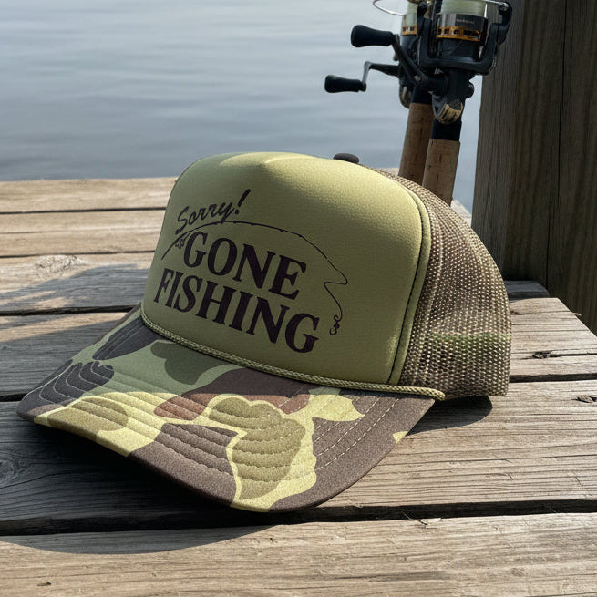 Camouflage fishing cap with 'Sorry! Gone Fishing' text sitting on a wooden dock next to fishing poles